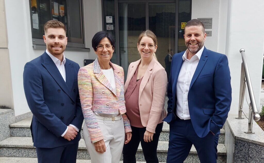 Vier Personen in Businesskleidung stehen lächelnd zusammen vor einem Gebäude mit Stufen und Glastüren