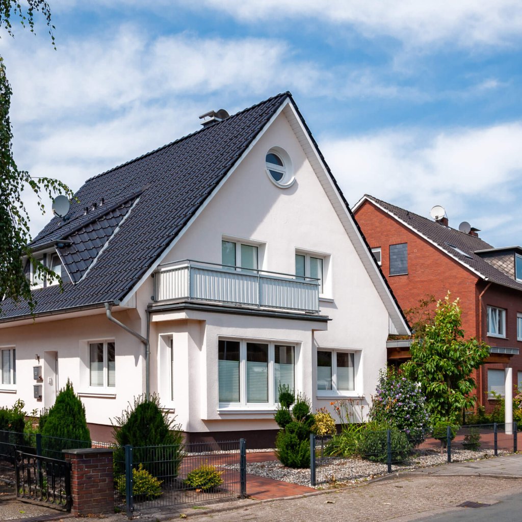 Ein weißes modernes Haus mit dunklem Dach, kleinem Garten und Gehweg steht in einem Wohnviertel unter blauem Himmel